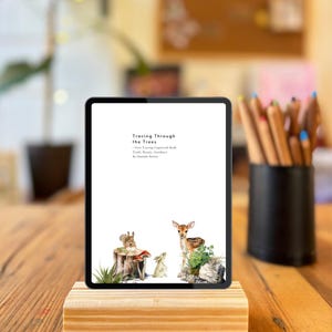 Pode incluir: Um tablet digital exibindo a capa do livro "Tracing Through the Trees". A capa apresenta uma ilustração em aquarela de um veado, um coelho e um esquilo em um cenário de floresta. O tablet repousa sobre um bloco de madeira.