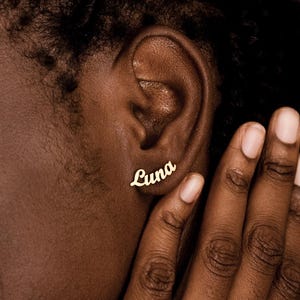 Può includere: Primo piano di un orecchino color oro che riporta il nome "Luna" in corsivo. L'orecchino è sull'orecchio di una persona con la pelle scura. La mano della persona è vicino all'orecchio.