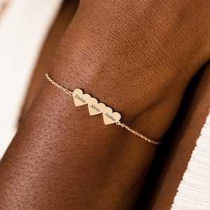 Puede incluir: Pulsera dorada con tres colgantes en forma de corazón grabados con los nombres Emma, Alexis y Amber. La pulsera se lleva en la muñeca. En el fondo, un suéter color crema.