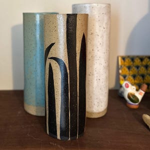 以下が含まれることがあります： さまざまな色とデザインの3つのセラミック花瓶。中央の花瓶は、黒い縦縞と葉のデザインが施されたタン色です。水色の花瓶と斑点のある白い花瓶が両側にあります。すべて円筒形です。