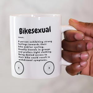 以下が含まれることがあります： 「Bikesexual」の黒い文字と定義、自転車の車輪のイラストが2つ描かれた白いセラミックマグ。マグカップは人が持っています。
