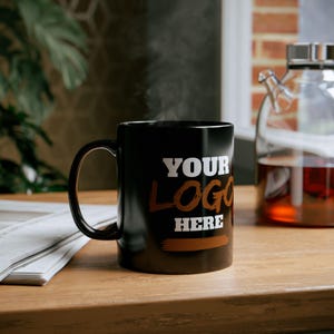 May include: A black ceramic mug with the text "YOUR LOGO HERE" in white and brown. The mug is on a wooden surface, with steam rising from it. A glass carafe with liquid is in the background.