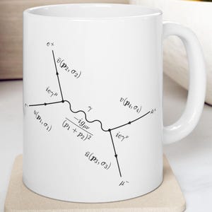 Può includere: Una tazza in ceramica bianca con un'illustrazione in bianco e nero di un diagramma di Feynman, una rappresentazione delle interazioni di particelle in fisica. Il diagramma mostra un'interazione di particelle con il testo 'ieγμ' e '-igμν/(p1+p2)2'.