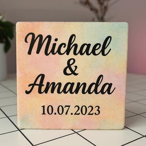 Puede incluir: Un bloque cuadrado con un fondo de estilo acuarela en colores pastel. Los nombres "Michael & Amanda" están escritos en una elegante escritura negra, con la fecha "10.07.2023" debajo. Un regalo personalizado para bodas o aniversarios.
