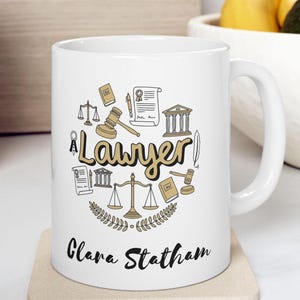 May include: White ceramic mug with a gold design of legal symbols and the word "Lawyer". The mug has the name "Clara Statham" printed on the bottom.