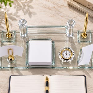 Puede incluir: Un conjunto organizador de escritorio de cristal transparente que incluye dos bolígrafos dorados, un soporte cuadrado para notas con papel blanco, un reloj analógico redondo con borde dorado y dos tarjeteros sobre una bandeja de cristal.