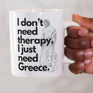 Pode incluir: Caneca de cerâmica branca com um gráfico preto de uma estátua grega e o texto "I don't need therapy, I just need Greece." A caneca tem uma alça curva.