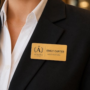 Puede incluir: Una placa de identificación rectangular de tono dorado con un acabado de metal cepillado. La placa muestra la letra A dentro de una corona de laurel, el texto AURORA HOTEL, el nombre EMILY CARTER y el cargo GUEST RELATIONS en letras negras.