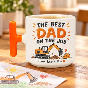 Könnte beinhalten: Ein weißer Keramikbecher mit einem leuchtend orangefarbenen T-förmigen Henkel. Auf der Vorderseite steht der Text "THE BEST DAD ON THE JOB" in Schwarz und Orange, eine orangefarbene Bagger-Illustration, ein kleiner orangefarbener Helm und "From Leo + Mia".