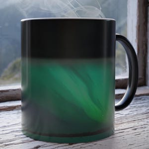 Puede incluir: Una taza de color cambiante negra y verde con vapor que sale de la parte superior. La taza está sobre una superficie de madera.