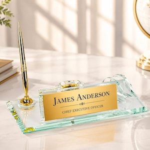 Puede incluir: Una placa de nombre para escritorio de cristal transparente con una placa metálica dorada que muestra el texto impreso "JAMES ANDERSON" y "CHIEF EXECUTIVE OFFICER" en negro. Incluye un bolígrafo dorado a juego en una base de cristal.