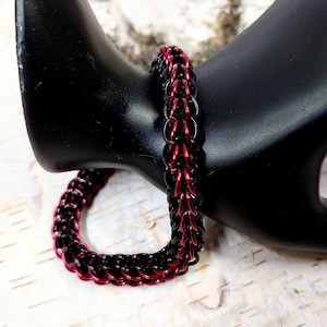 May include: A handmade chainmail bracelet with alternating black and red rings. The bracelet is displayed on a black mannequin hand, showcasing its intricate weave and color contrast. The jewelry piece is a striking accessory.