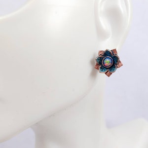 May include: A pair of copper earrings with a blue and green flower design and an iridescent opal gemstone in the center.