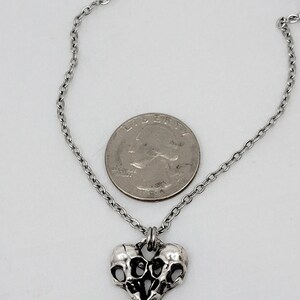 Puede incluir: Un collar de plata con un colgante en forma de coraz&oacute;n que presenta dos calaveras entrelazadas. El colgante es aproximadamente del tama&ntilde;o de una moneda de 25 centavos.