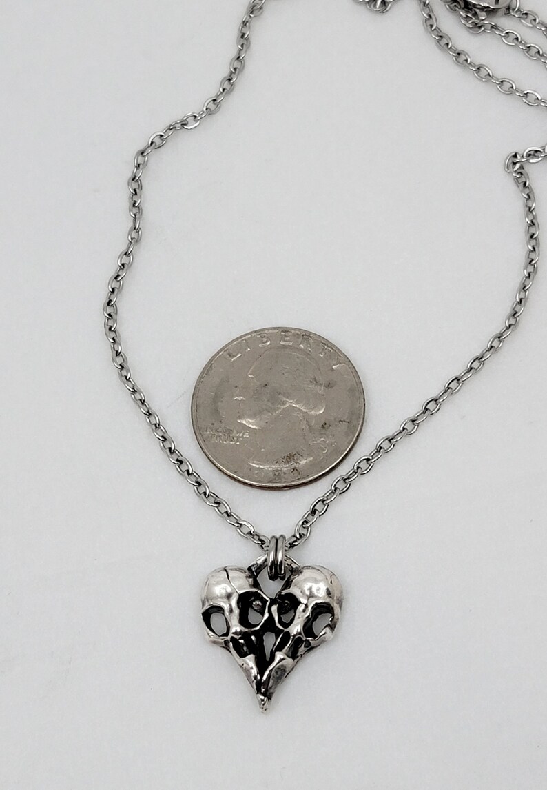 Puede incluir: Un collar de plata con un colgante en forma de coraz&oacute;n que presenta dos calaveras entrelazadas. El colgante es aproximadamente del tama&ntilde;o de una moneda de 25 centavos.