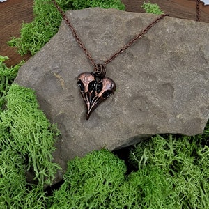 Puede incluir: Un collar de cobre con un colgante en forma de coraz&oacute;n que presenta tres cr&aacute;neos de p&aacute;jaros estilizados. El colgante es detallado y tiene un acabado de p&aacute;tina oscura.