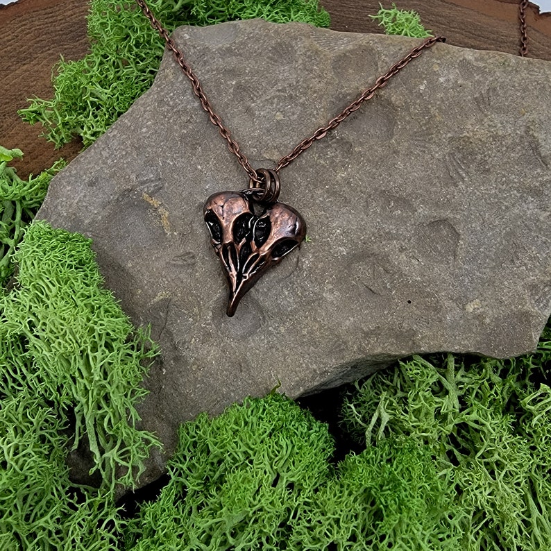 Puede incluir: Un collar de cobre con un colgante en forma de coraz&oacute;n que presenta tres cr&aacute;neos de p&aacute;jaros estilizados. El colgante es detallado y tiene un acabado de p&aacute;tina oscura.