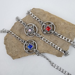 May include: Three silver chain bracelets with a circular design featuring a different coloured gemstone in each. The gemstones are red, blue, and iridescent.