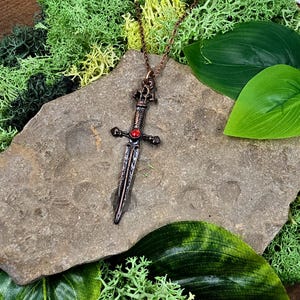 May include: A copper-toned sword pendant with a red gemstone set in the hilt. The pendant is hanging from a copper chain on a brown stone background.