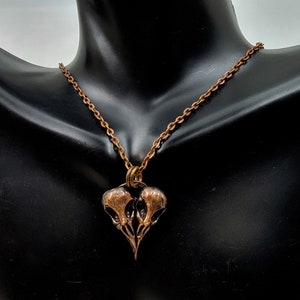 Puede incluir: Un collar de cobre con un colgante que presenta dos cr&aacute;neos de p&aacute;jaros estilizados. El colgante tiene forma de coraz&oacute;n.
