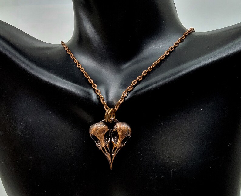 Puede incluir: Un collar de cobre con un colgante que presenta dos cr&aacute;neos de p&aacute;jaros estilizados. El colgante tiene forma de coraz&oacute;n.