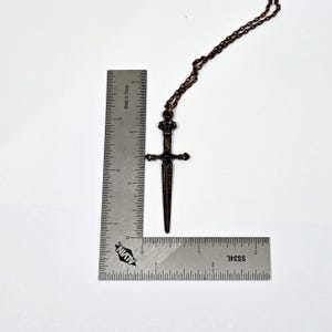 May include: A copper-toned sword pendant necklace with a black gemstone at the hilt. The pendant is approximately 5 cm long and hangs from a copper chain.