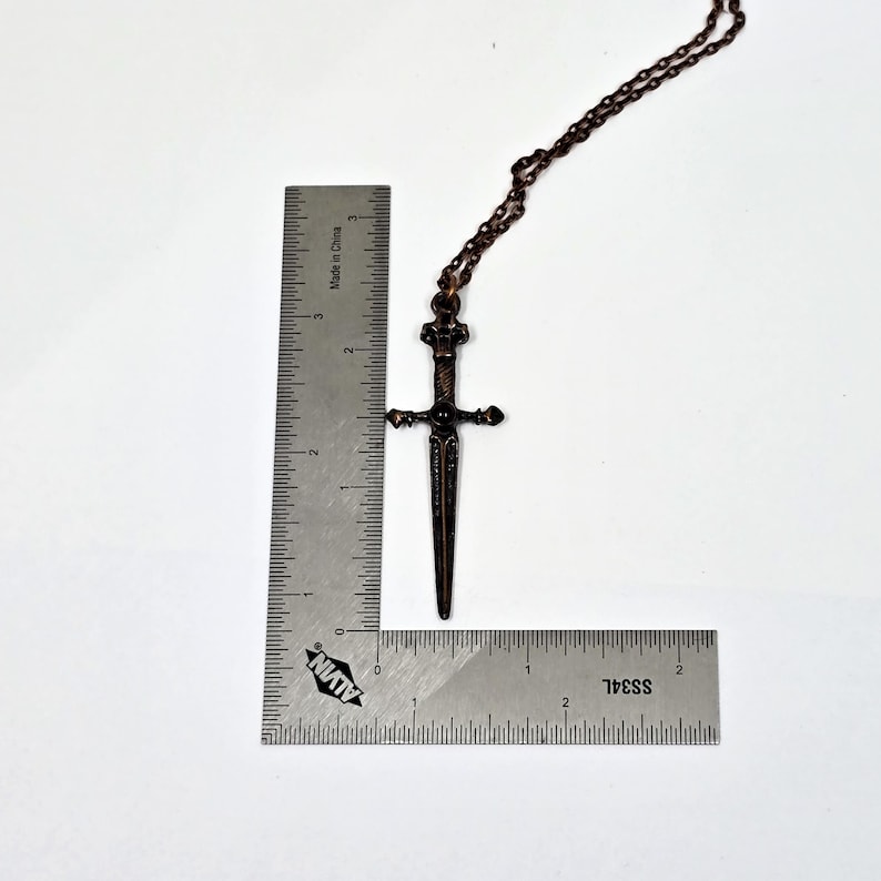 May include: A copper-toned sword pendant necklace with a black gemstone at the hilt. The pendant is approximately 5 cm long and hangs from a copper chain.