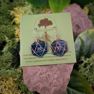 Puede incluir: Un par de pendientes de cobre y turquesa azul con forma de dado de veinte caras. Los pendientes están en una tarjeta verde con el texto "Tangled Earth Arts" y las manijas de las redes sociales.