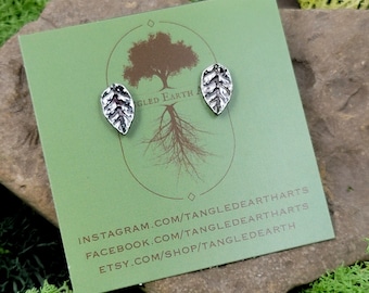 Puede incluir: Un par de pendientes de hoja de plata en una tarjeta verde con el texto "Tangled Earth" y los identificadores de redes sociales para Instagram, Facebook y Etsy.