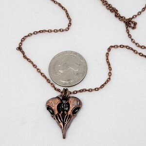 Puede incluir: Un collar de cobre con un colgante de calavera de p&aacute;jaro detallado. El colgante tiene forma de coraz&oacute;n y un acabado desgastado. El collar se muestra junto a una moneda de 20 c&eacute;ntimos de euro para referencia de tama&ntilde;o.