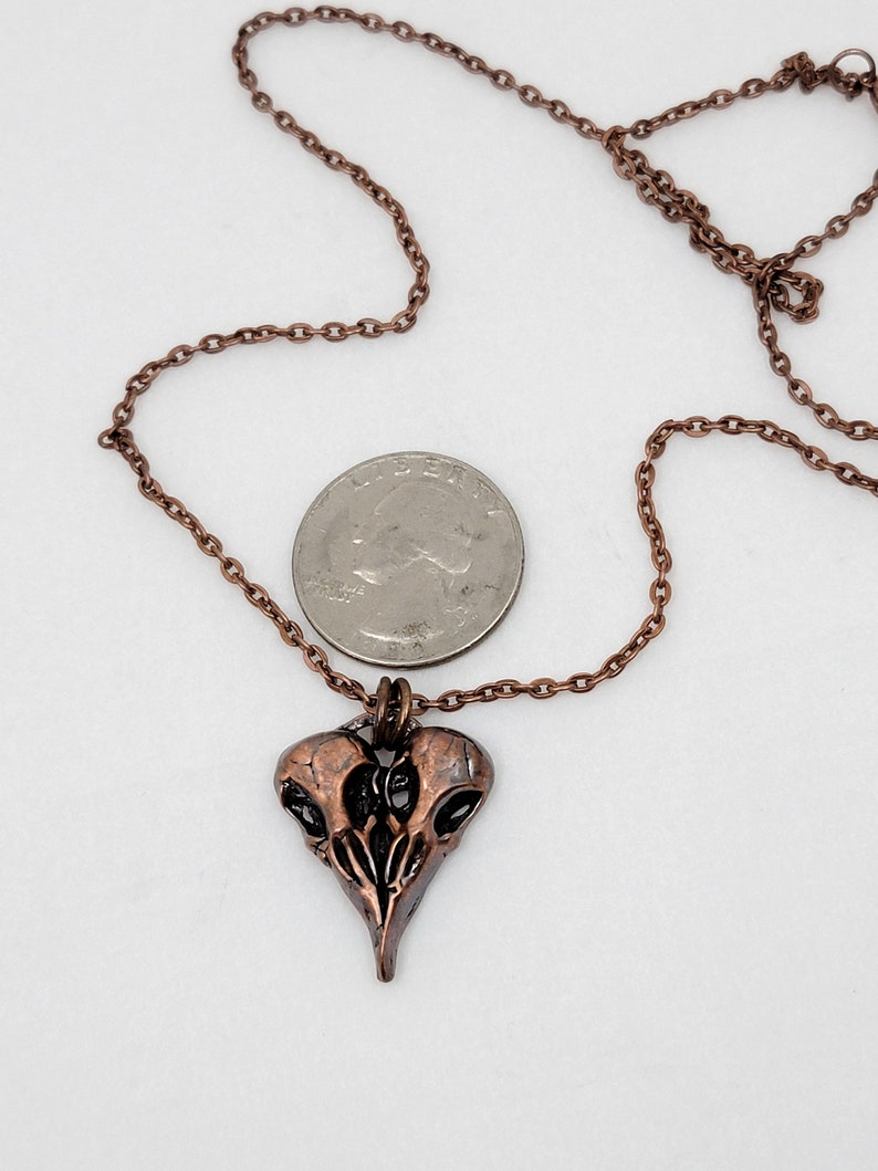 Puede incluir: Un collar de cobre con un colgante de calavera de p&aacute;jaro detallado. El colgante tiene forma de coraz&oacute;n y un acabado desgastado. El collar se muestra junto a una moneda de 20 c&eacute;ntimos de euro para referencia de tama&ntilde;o.