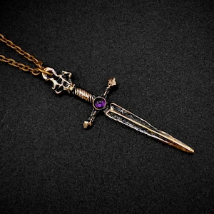Puede incluir: Un colgante de espada de color dorado con una piedra preciosa púrpura engastada en la empuñadura. La espada tiene un diseño detallado y está unida a una cadena dorada.