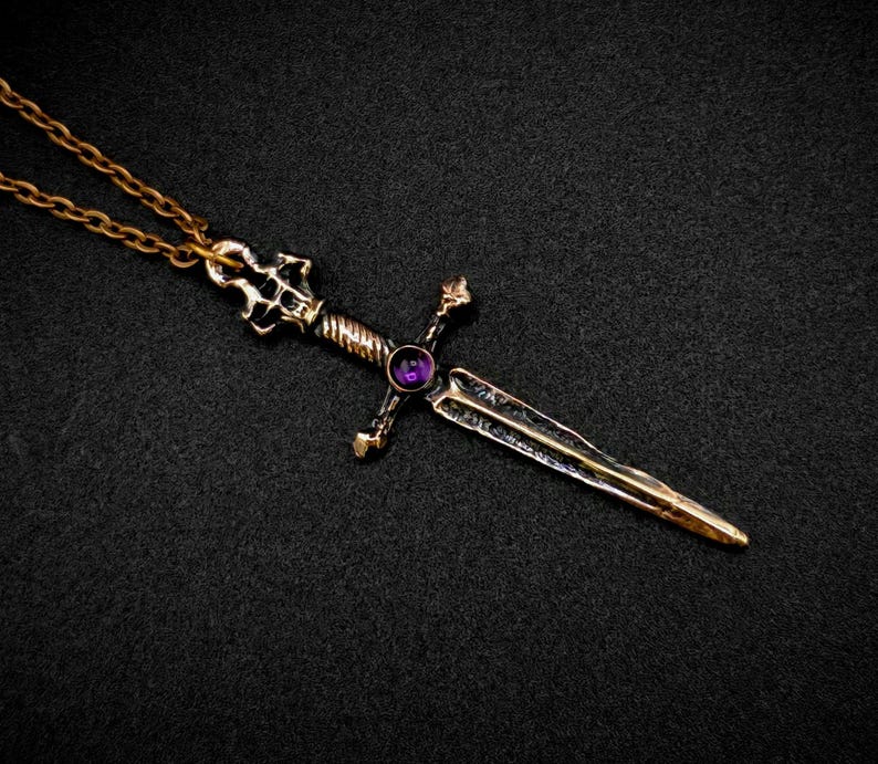 May include: A gold-toned sword pendant with a purple gemstone set in the hilt. The sword has a detailed design and is attached to a gold chain.