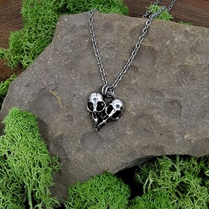 Puede incluir: Un collar de plata con un colgante de dos cr&aacute;neos de p&aacute;jaro unidos en forma de coraz&oacute;n. El collar est&aacute; sobre una roca gris con musgo verde.