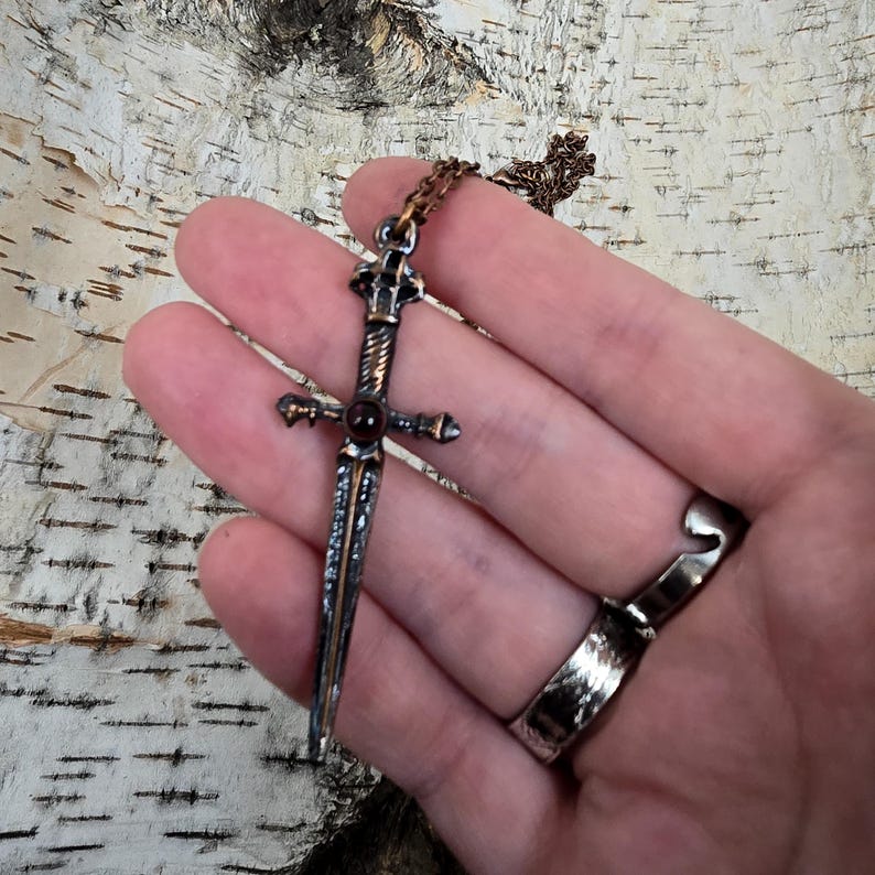 May include: A silver and copper sword pendant with a red gemstone in the centre. The pendant is on a silver chain.
