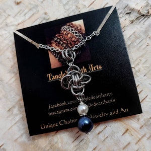 May include: A silver chain necklace with a knot pendant and two pearls, one white and one black. The necklace is displayed on a black card with the text "Tangle Earth Arts" and contact information. Unique Chain Jewelry and Art.
