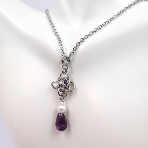 May include: A silver chain necklace with a silver knot pendant and a purple teardrop-shaped gemstone hanging from a white pearl.