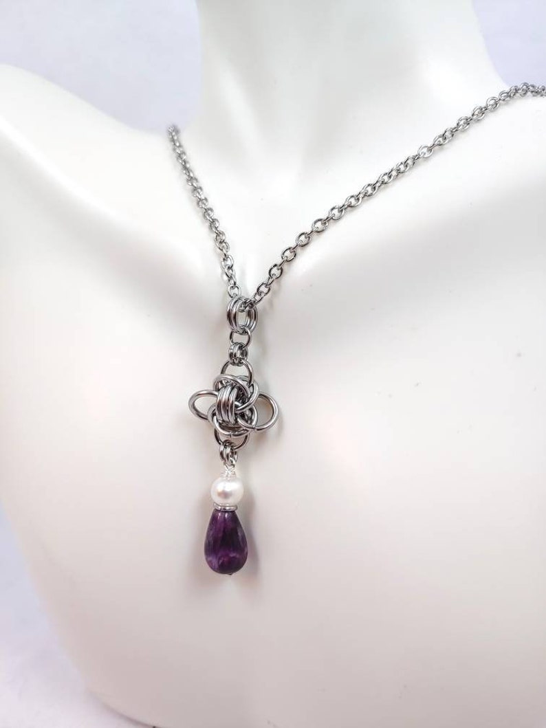 May include: A silver chain necklace with a silver knot pendant and a purple teardrop-shaped gemstone hanging from a white pearl.