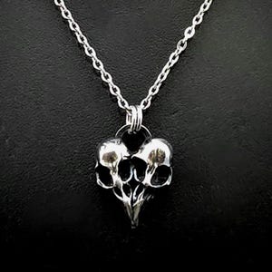 Puede incluir: Un collar de plata con un colgante en forma de coraz&oacute;n con dos calaveras entrelazadas.