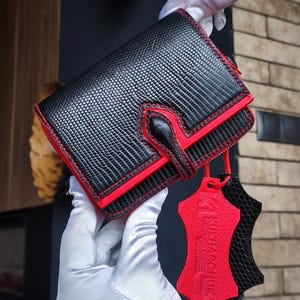 Puede incluir: Cartera de cuero negro y rojo con un patrón texturizado y costuras rojas. La cartera tiene un cierre de solapa y una pequeña etiqueta roja con texto. La cartera es sostenida por una persona que usa guantes blancos.