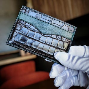 Puede incluir: Un tarjetero rectangular con una textura oscura y brillante de piel de cocodrilo en relieve. El accesorio incluye una banda horizontal de cuero liso y mate en color verde azulado en la parte superior, con costuras visibles en los bordes.