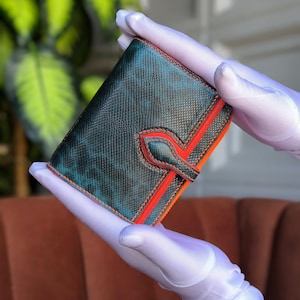 Op de afbeelding: Een teal en zwarte leren portemonnee met oranje randen en stiksels. De portemonnee heeft een textuur en een klepsluiting met een decoratief accent. De portemonnee wordt vastgehouden door een persoon met witte handschoenen.