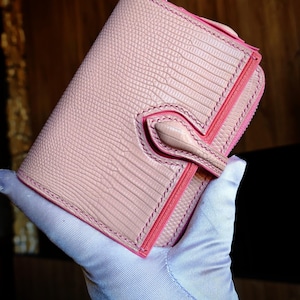 Peut inclure: Un portefeuille pliable rose clair avec un motif texturé en peau de lézard. Le portefeuille présente une fermeture à languette centrale, des coutures visibles sur les bords et une doublure intérieure rose vif visible sur la couture latérale.