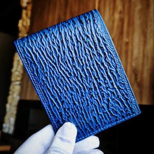 Puede incluir: Una cartera rectangular azul con una superficie de textura profunda y rugosa que recuerda a la corteza de un árbol o a la piedra erosionada. La cartera tiene bordes cosidos limpios y un color azul cobalto vibrante y uniforme.