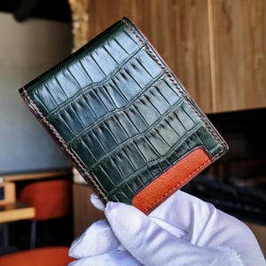 May include: A rectangular wallet featuring a dark green crocodile-embossed leather exterior with a glossy finish. The wallet includes a contrasting tan leather accent panel at the bottom corner and is constructed with visible orange stitching along the edges.