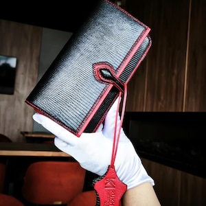 Könnte beinhalten: Schwarze Ledergeldbörse mit roter Umrandung und Nähten. Die Geldbörse hat eine strukturierte Oberfläche und ein rotes Lederetikett mit dem Markennamen "KUKHARCHUK". Die Geldbörse wird von einer behandschuhten Hand gehalten.