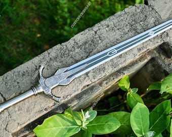 May include: A silver and black sword with a decorative handle and a long, pointed blade. The blade has a central black stripe with a geometric design. The sword is resting on a weathered stone surface, surrounded by green foliage.
