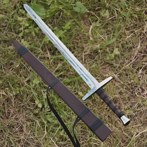 Puede incluir: Una espada plateada con guarda y empuñadura de cuero marrón descansa sobre la hierba junto a una vaina de cuero marrón. La hoja de la espada es larga y puntiaguda, y la vaina tiene una correa negra.