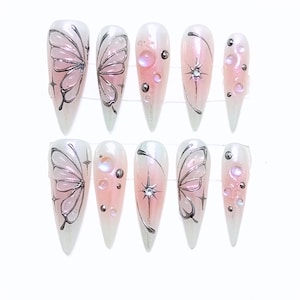 Puede incluir: Un conjunto de diez uñas postizas largas y puntiagudas. Cada uña presenta un diseño degradado, que transiciona de blanco a rosa. Algunas uñas tienen diseños de alas de mariposa, otras tienen estrellas y adornos iridiscentes.