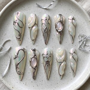 May include: A set of ten almond-shaped press-on nails featuring a pearlescent white base with raised silver metallic accents, intricate swirling lines, and embedded oval-shaped light pink stones on select nails, arranged on a speckled plate.